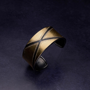 A gold bangle crossed with a wide black "X" and outlined with small rough diamonds floats above a deep grey backdrop.